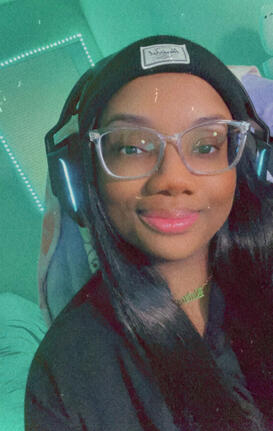 a selfie of mira. a black, femme presenting nonbinary person. they are wearing a black beanie, clear frame glasses, a black t-shirt, and a gold chain. the room is illuminated by strip lights in the background.