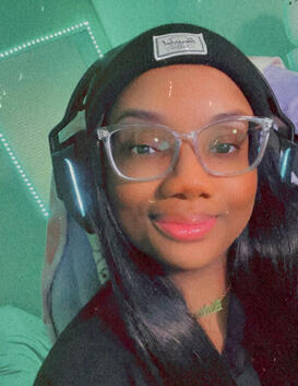 a selfie of mira. a black, femme presenting nonbinary person. they are wearing a black beanie, clear frame glasses, a black t-shirt, and a gold chain. the room is illuminated by strip lights in the background.